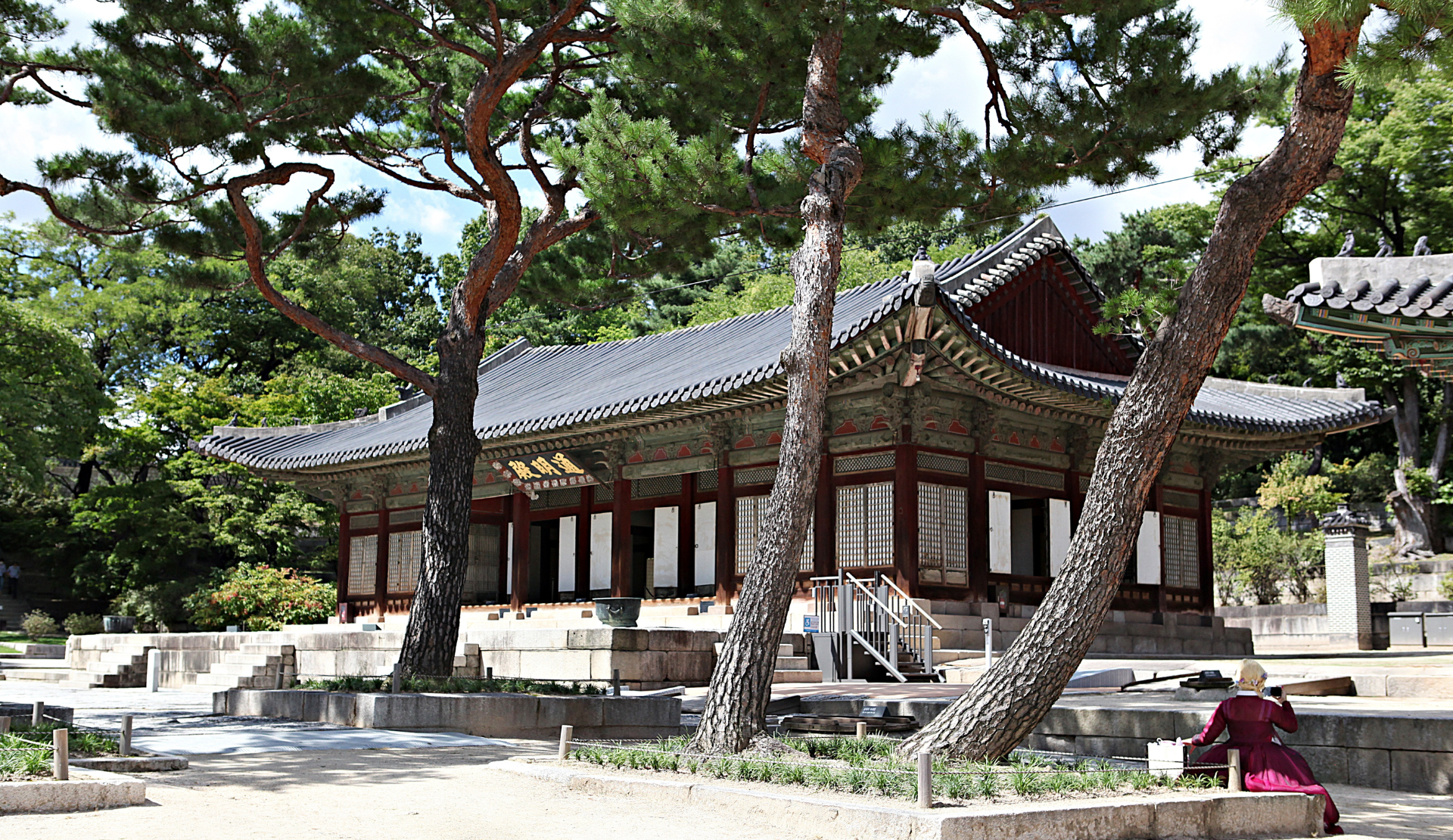 Blick auf ein Palastgebäude des Changgyeonggung unter alten Kiefern in Seoul, im Vordergrund eine Frau in traditionellem Hanbok
