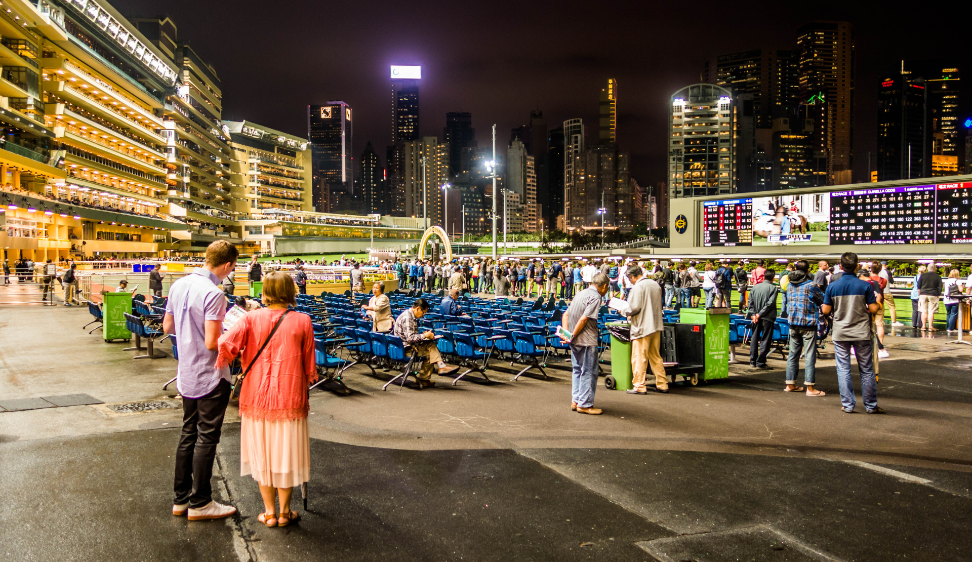 Paar beobachtet Vorbereitungen bei Happy Valley Racecourse in Hongkong mit aufgestellten Stühlen und beleuchteter Skyline bei Nacht