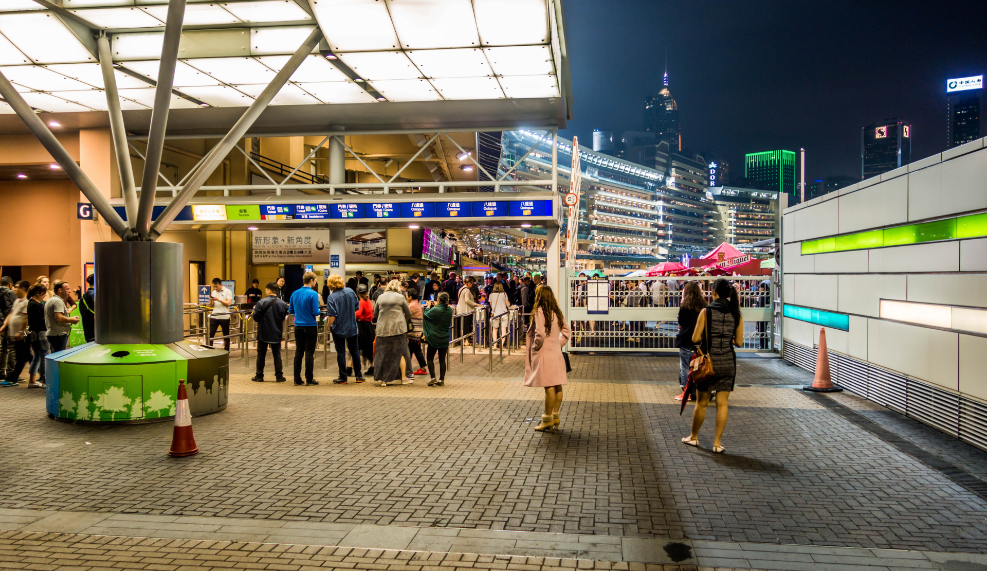 Moderner Eingangsbereich mit Wettschaltern bei Happy Valley Racecourse in Hongkong mit Besucherschlangen und beleuchteter Skyline im Hintergrund