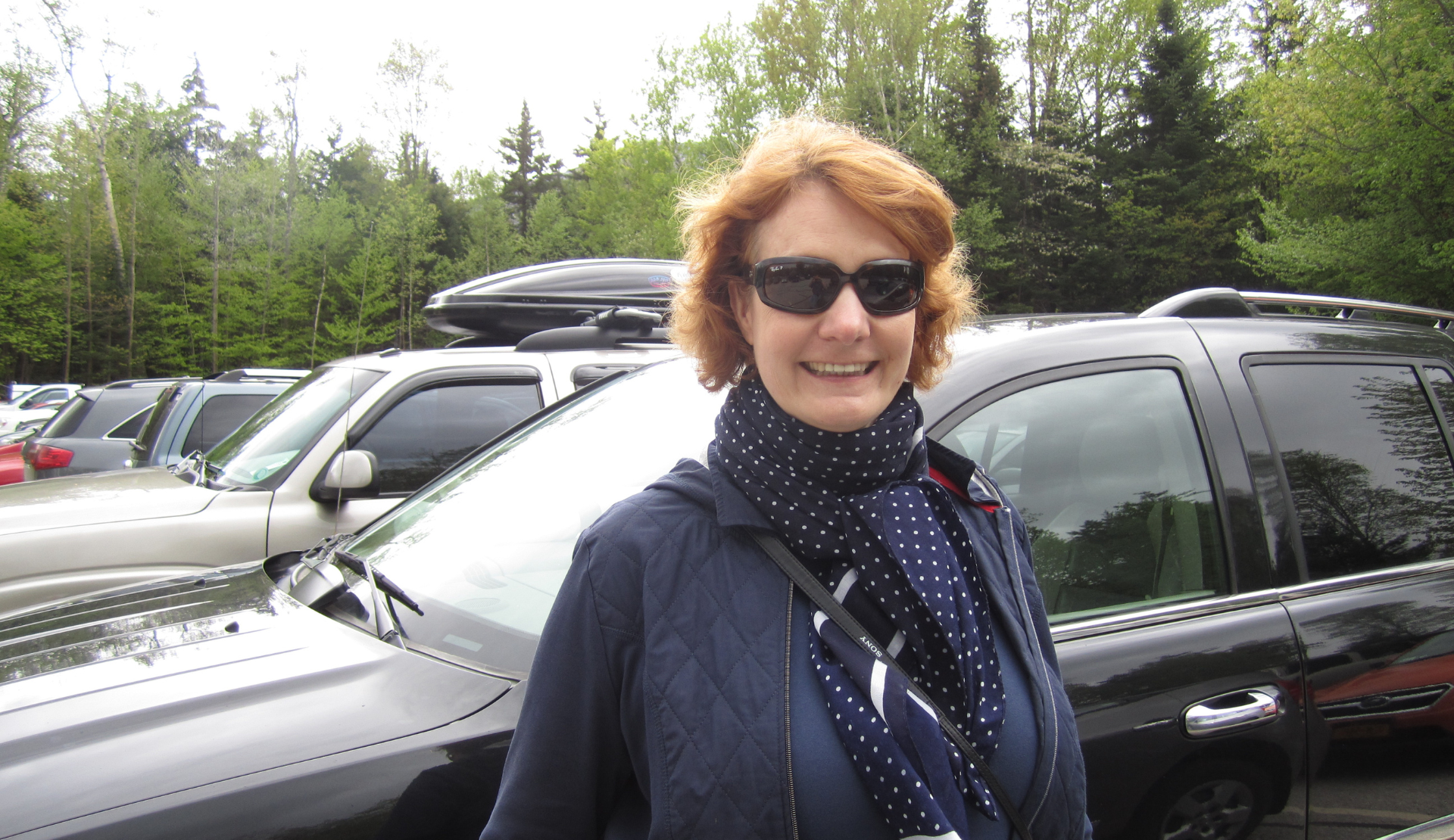 Peggy Zöllner steht lächelnd auf einem Parkplatz vor geparkten Autos und Bäumen im Hintergrund