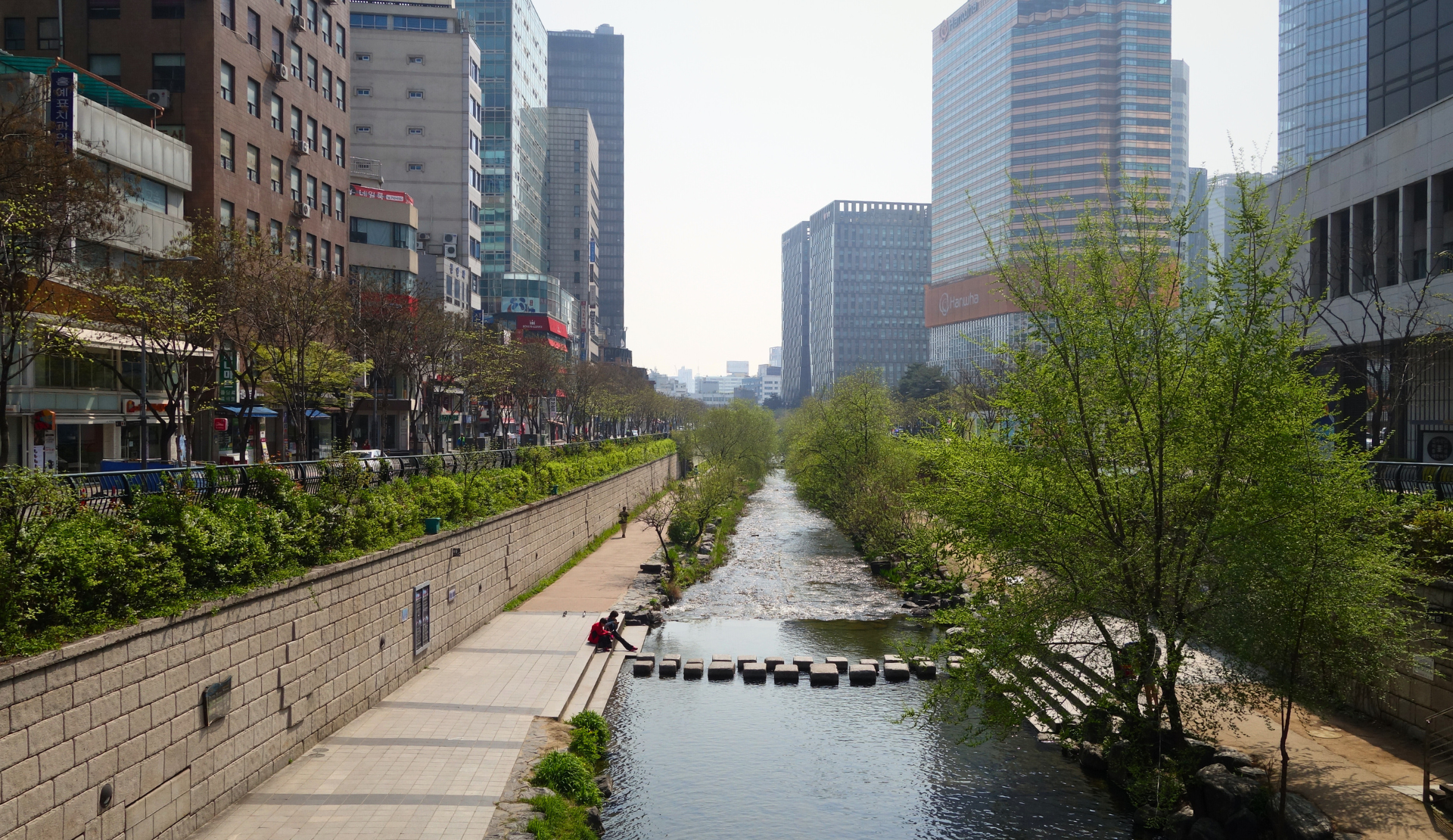 Blick entlang des Cheonggyecheon-Streams in Seoul mit Spazierwegen, grüner Vegetation und Hochhäusern im Hintergrund