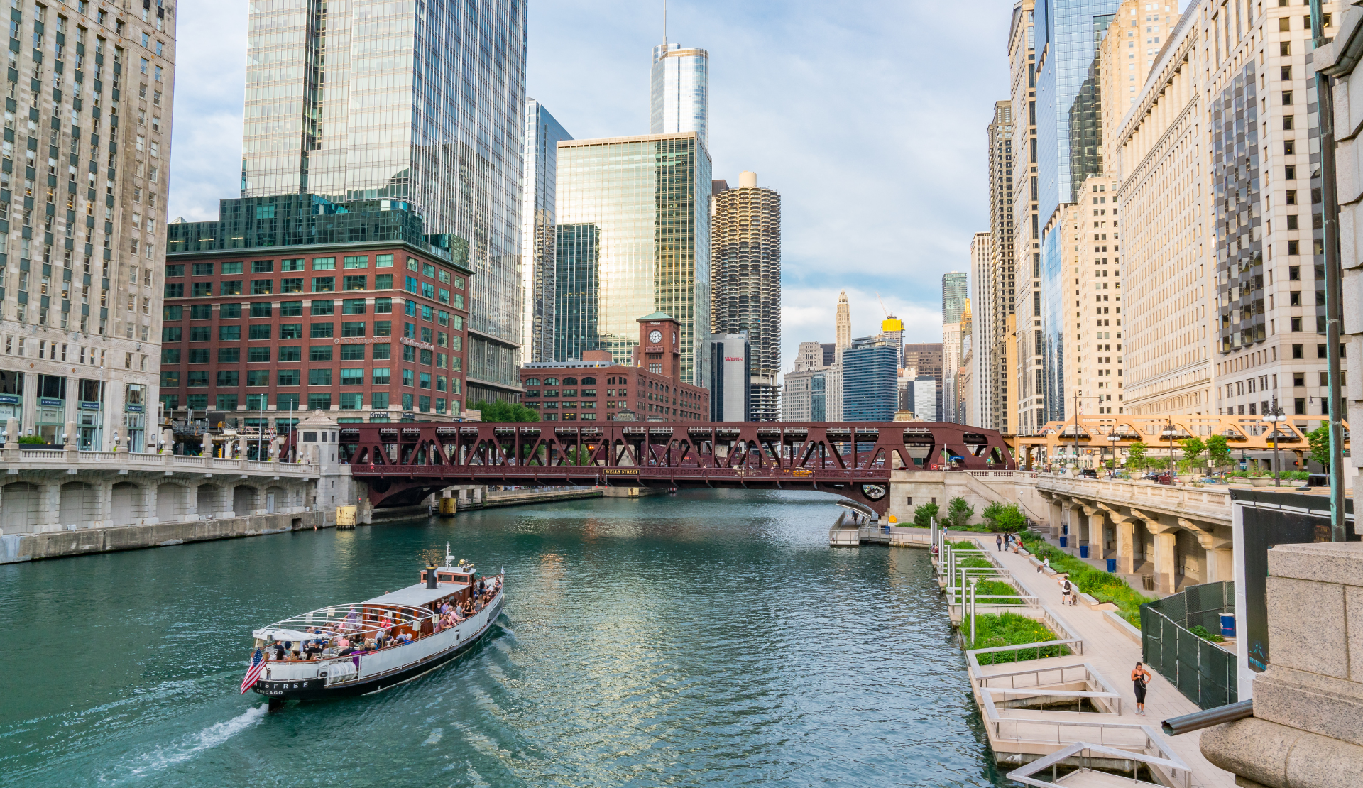Der Chicago River mit Ausflugsboot, dem Riverwalk am Ufer und der Skyline von Downtown Chicago mit Wolkenkratzern