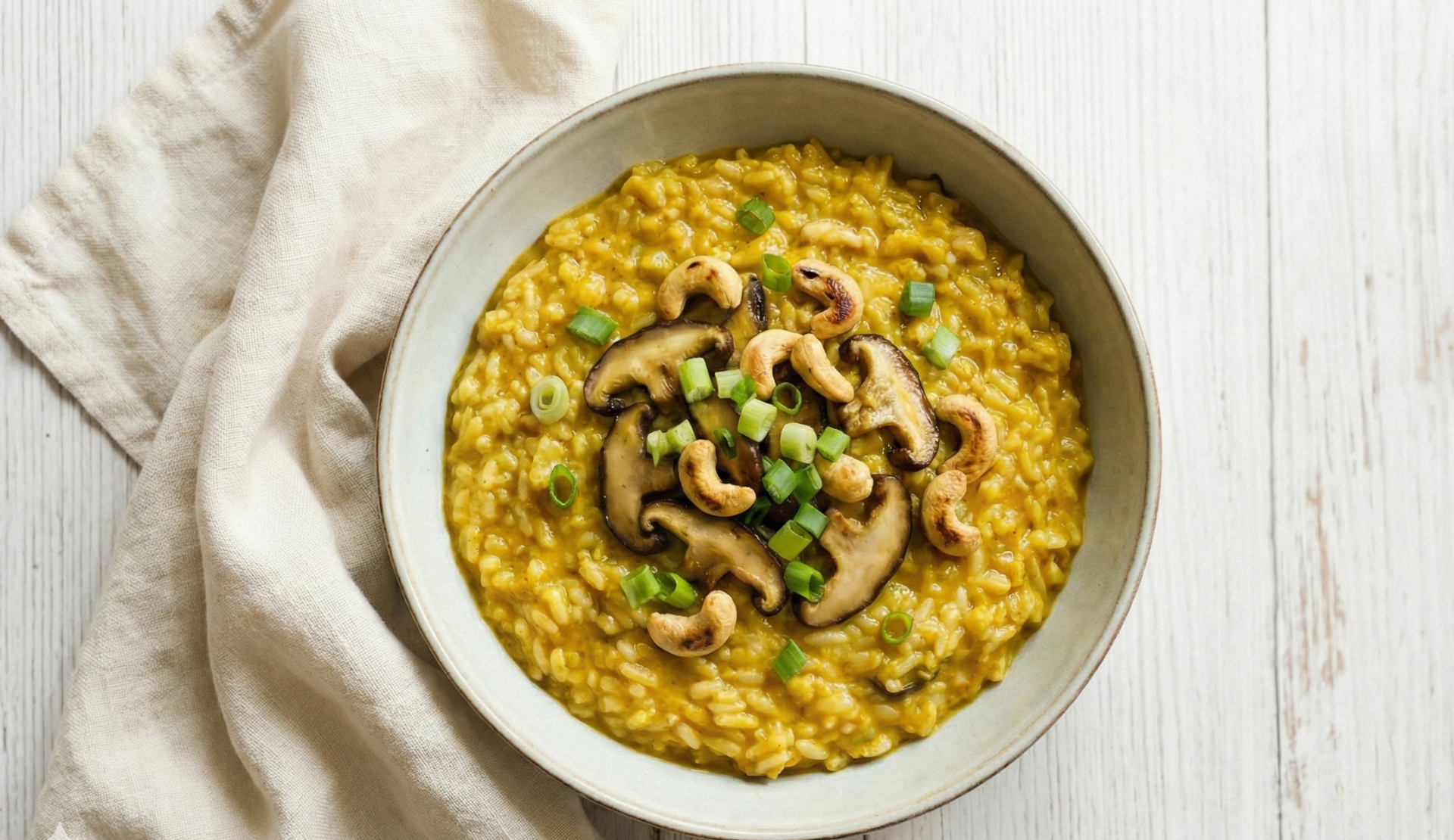 Nahaufnahme von Curry-Risotto mit Shiitake-Pilzen, Cashews und Frühlingszwiebeln in weißer Schüssel auf hellem Holztisch
