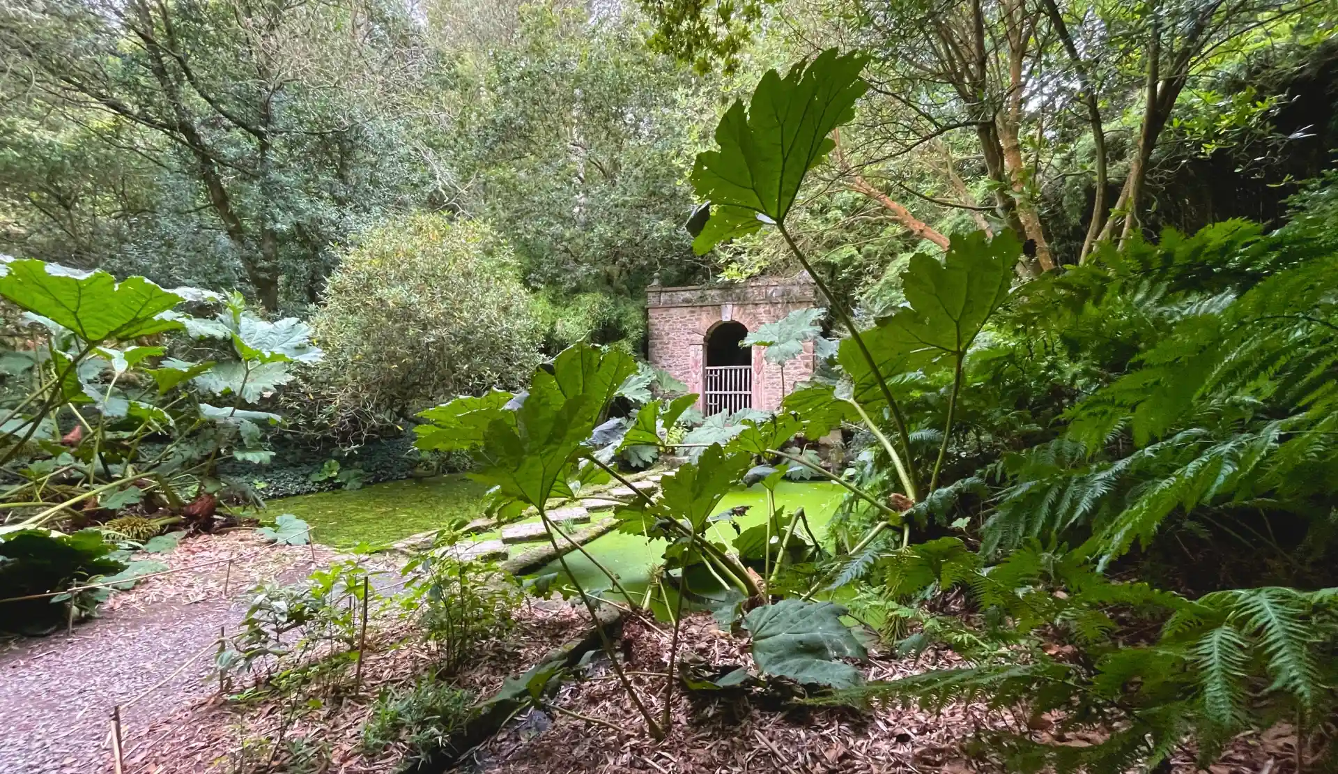 Verwunschener Gartenweg mit üppiger Vegetation und Torbogen in den Gärten von Kerdalo