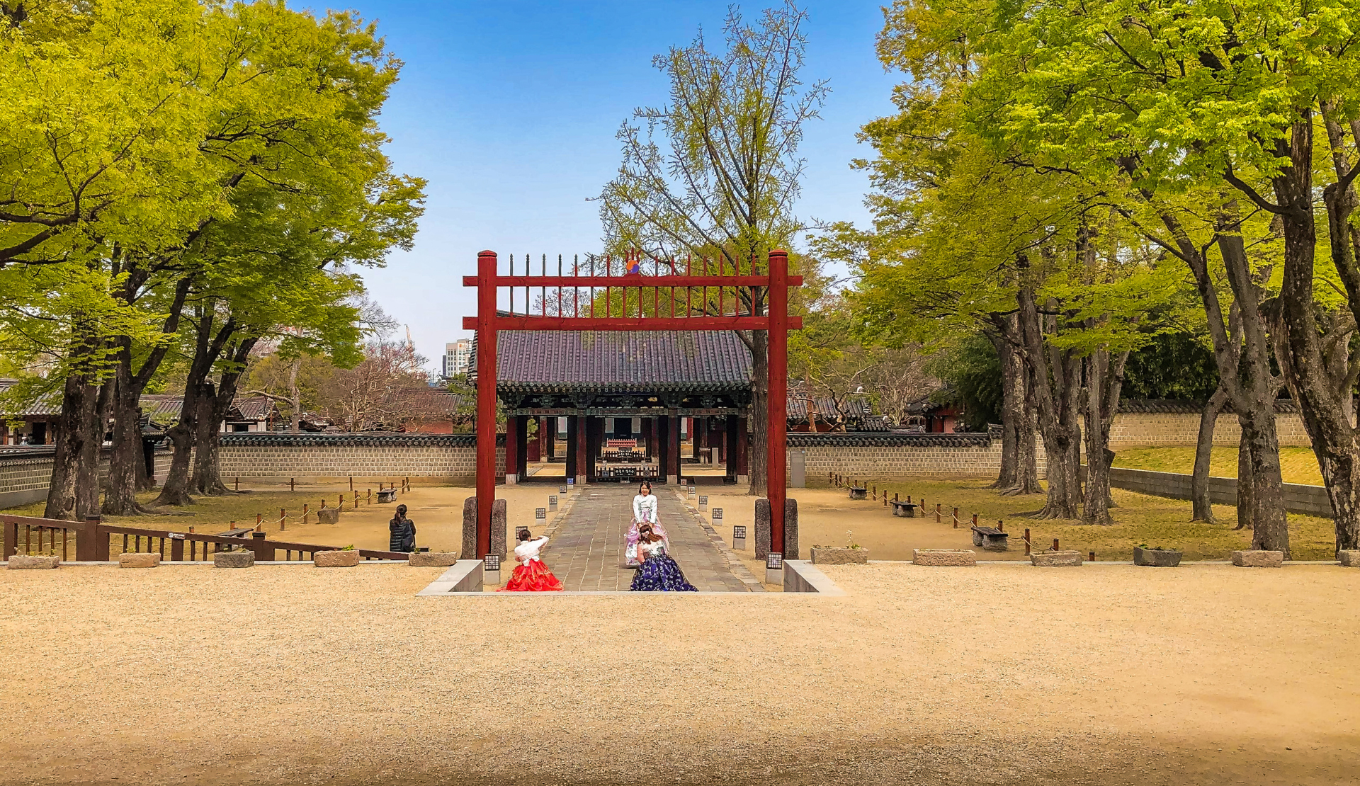Blick auf das rote Eingangstor des Gyeonggijeon-Schreins in Jeonju mit alten Bäumen und Besuchern in Hanbok
