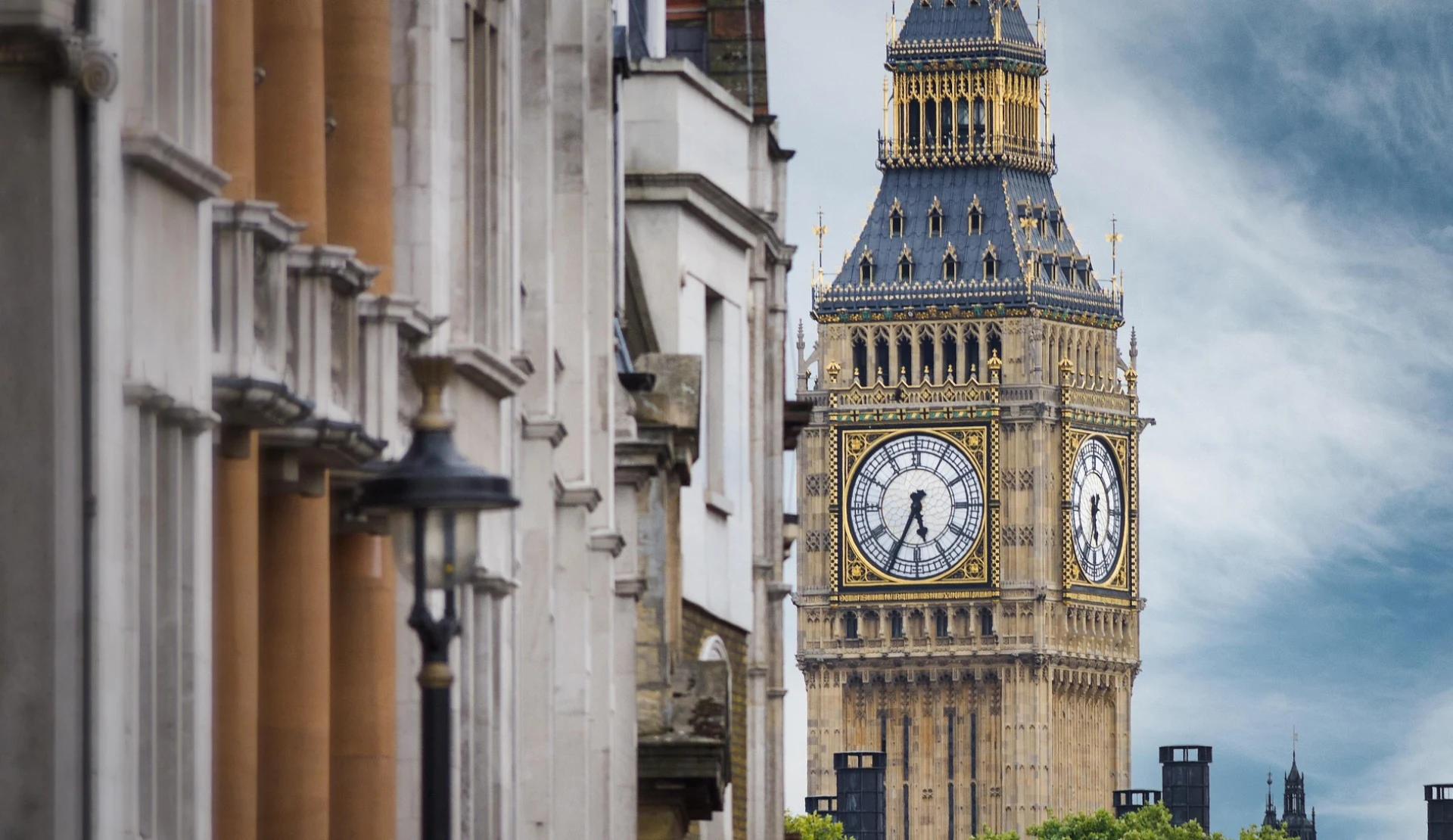 Der Glockenturm Big Ben in London mit goldverzierter Turmuhr und Spitze, links im Bild helle Häuserfassaden