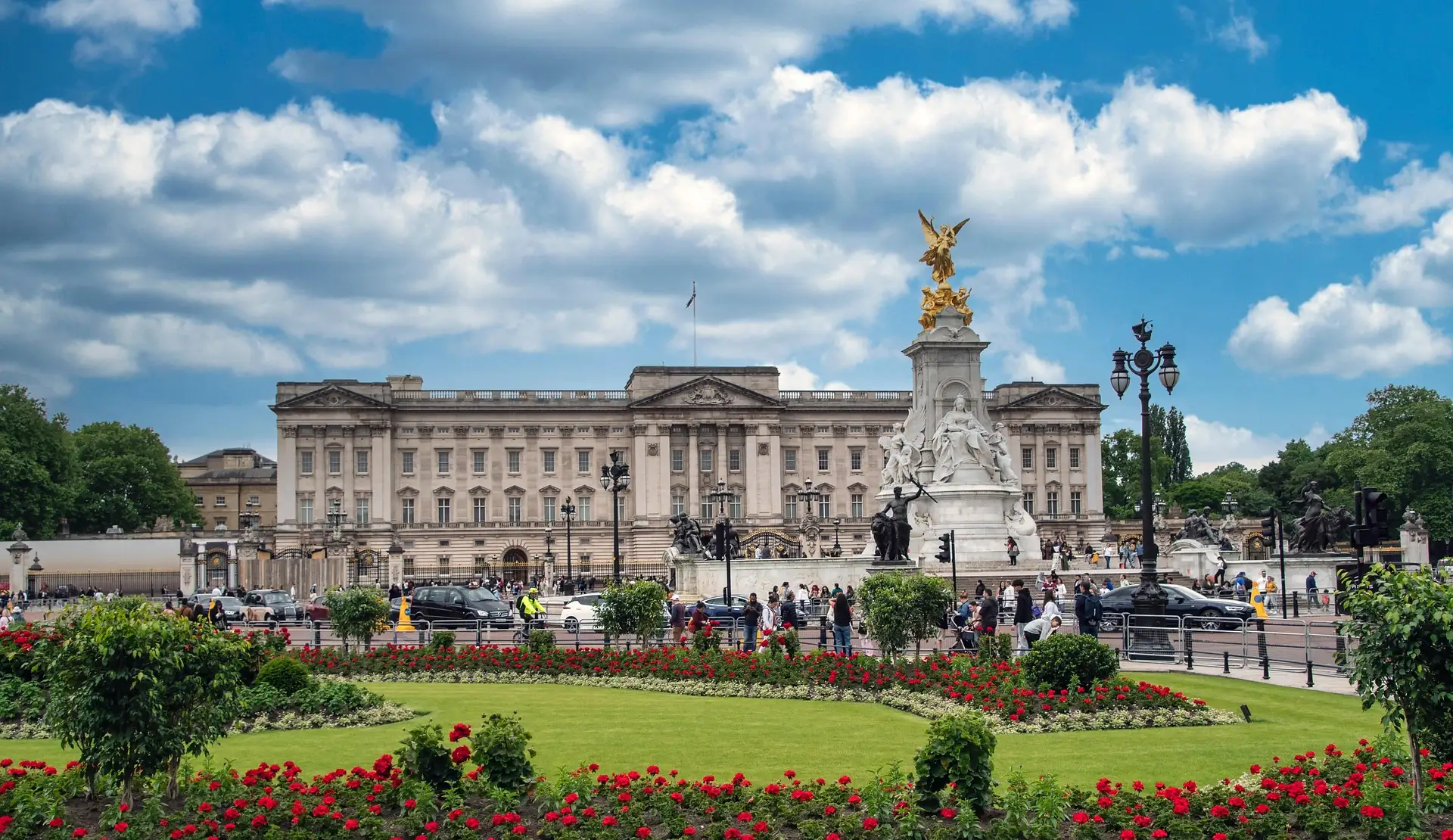 Buckingham Palace mit dem Victoria Memorial, davor Rasenflächen mit roten Blumenbeeten unter bewölktem Himmel