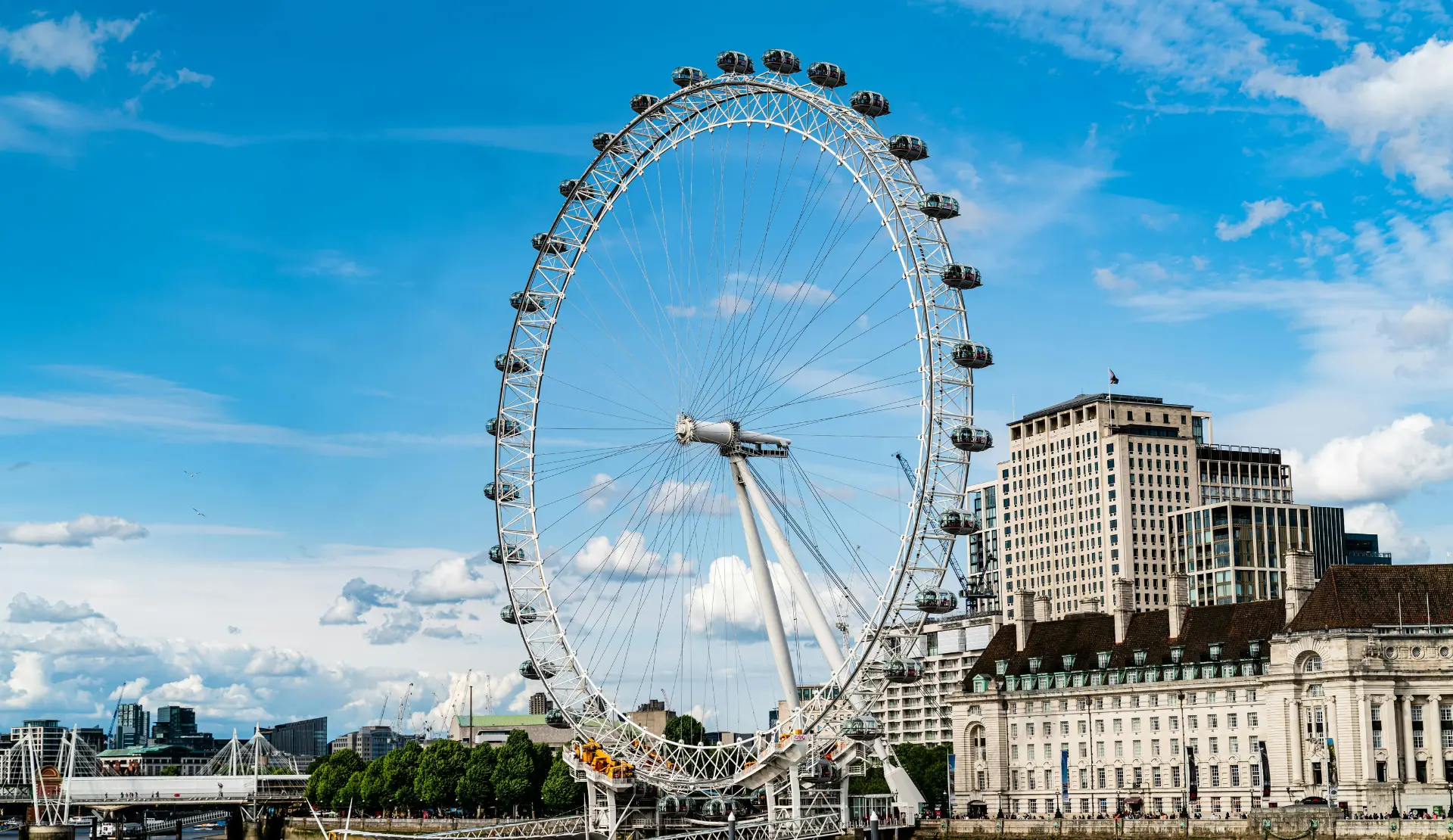 Das London Eye in Nahaufnahme mit weißen Speichen und Gondeln vor blauem Himmel, rechts ein historisches Gebäude am Ufer