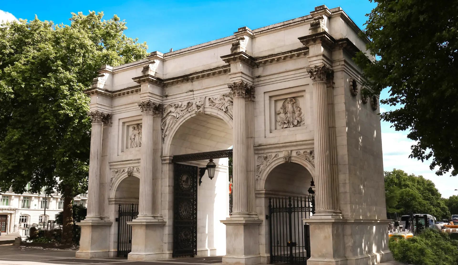 Der Marble Arch in London, ein heller Triumphbogen mit Säulen und schwarzen Eisentoren vor Bäumen und blauem Himmel