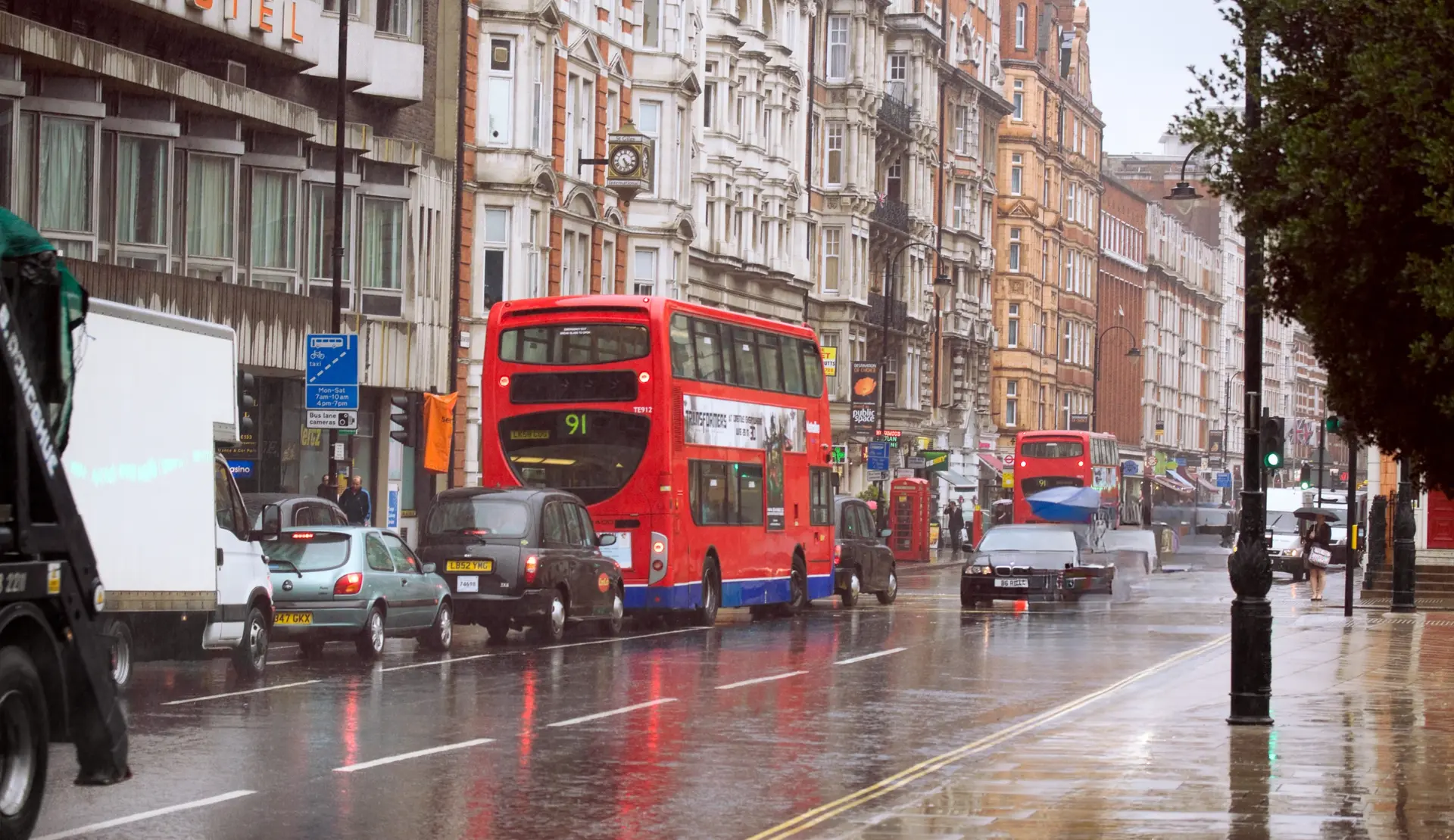 Regnerische Straße in London mit roten Doppeldeckerbussen, einem schwarzen Taxi und nassem Asphalt vor hohen Häusern