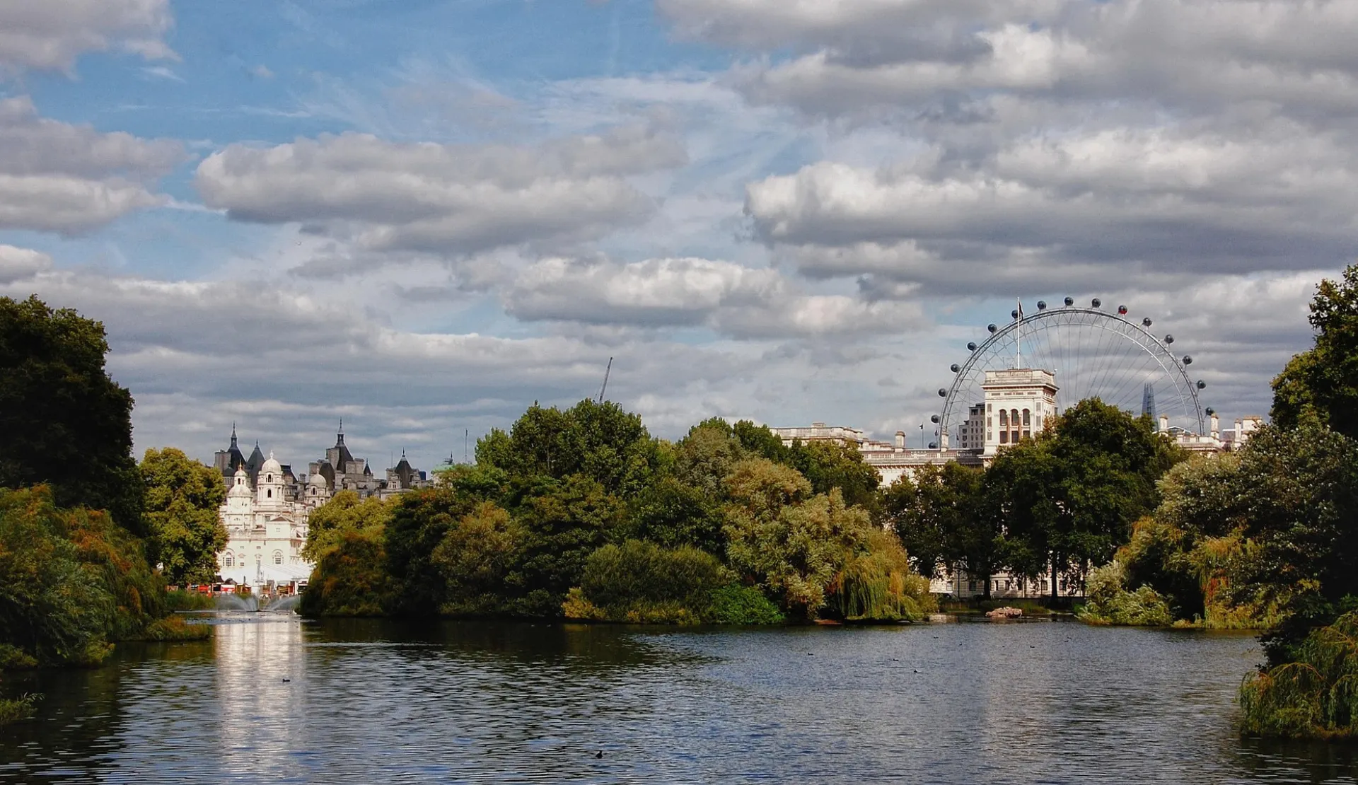 Blick über den See im St. James's Park in London mit Bäumen, hellen Gebäuden am Ufer und dem London Eye im Hintergrund