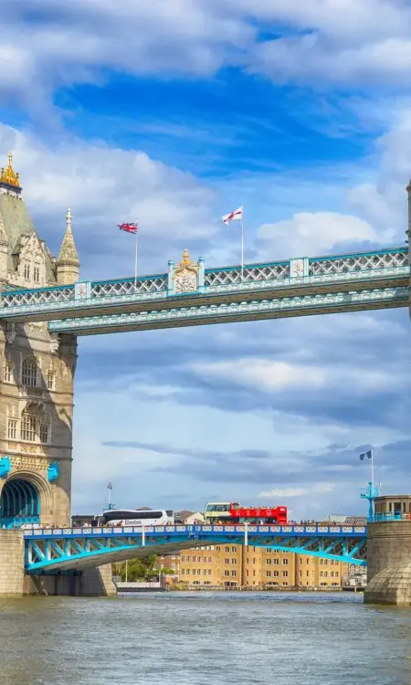Die Tower Bridge in London mit ihren zwei Türmen und der türkisfarbenen Stahlkonstruktion vor bewölktem Himmel, davor die Themse