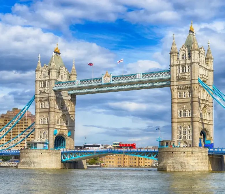 Die Tower Bridge in London mit ihren zwei Türmen und der türkisfarbenen Stahlkonstruktion vor bewölktem Himmel, davor die Themse