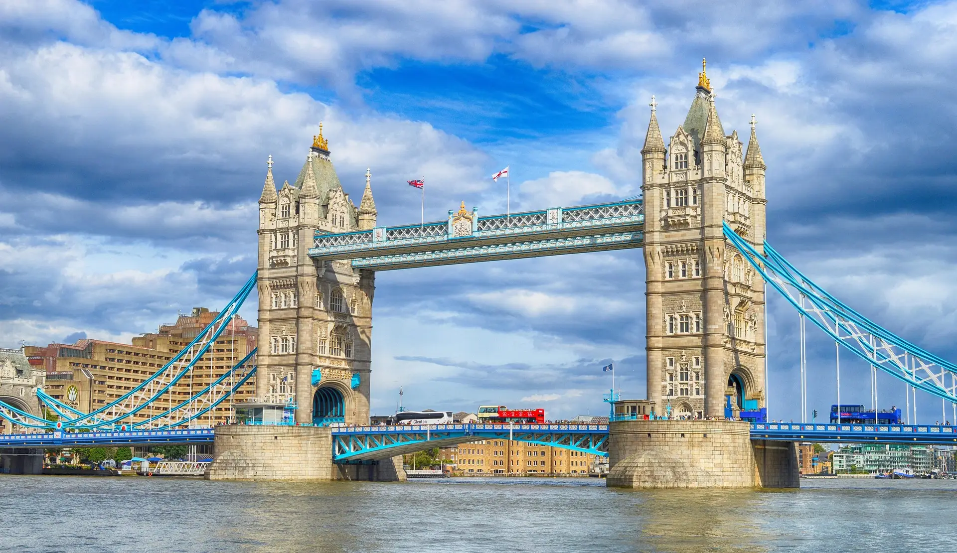 Die Tower Bridge in London mit ihren zwei Türmen und der türkisfarbenen Stahlkonstruktion vor bewölktem Himmel, davor die Themse
