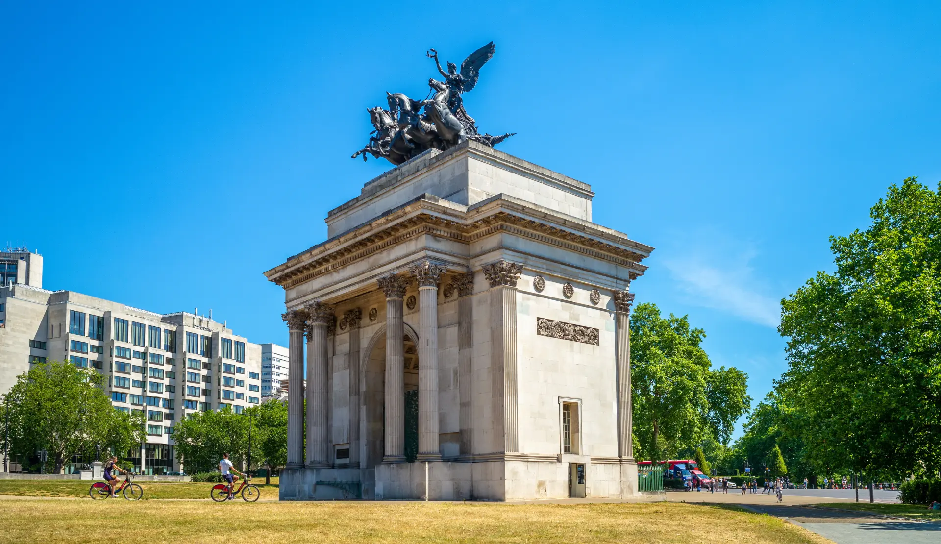 Der Wellington Arch in London mit Bronzeskulptur und Pferdegespann auf der Spitze, davor zwei Radfahrer unter blauem Himmel