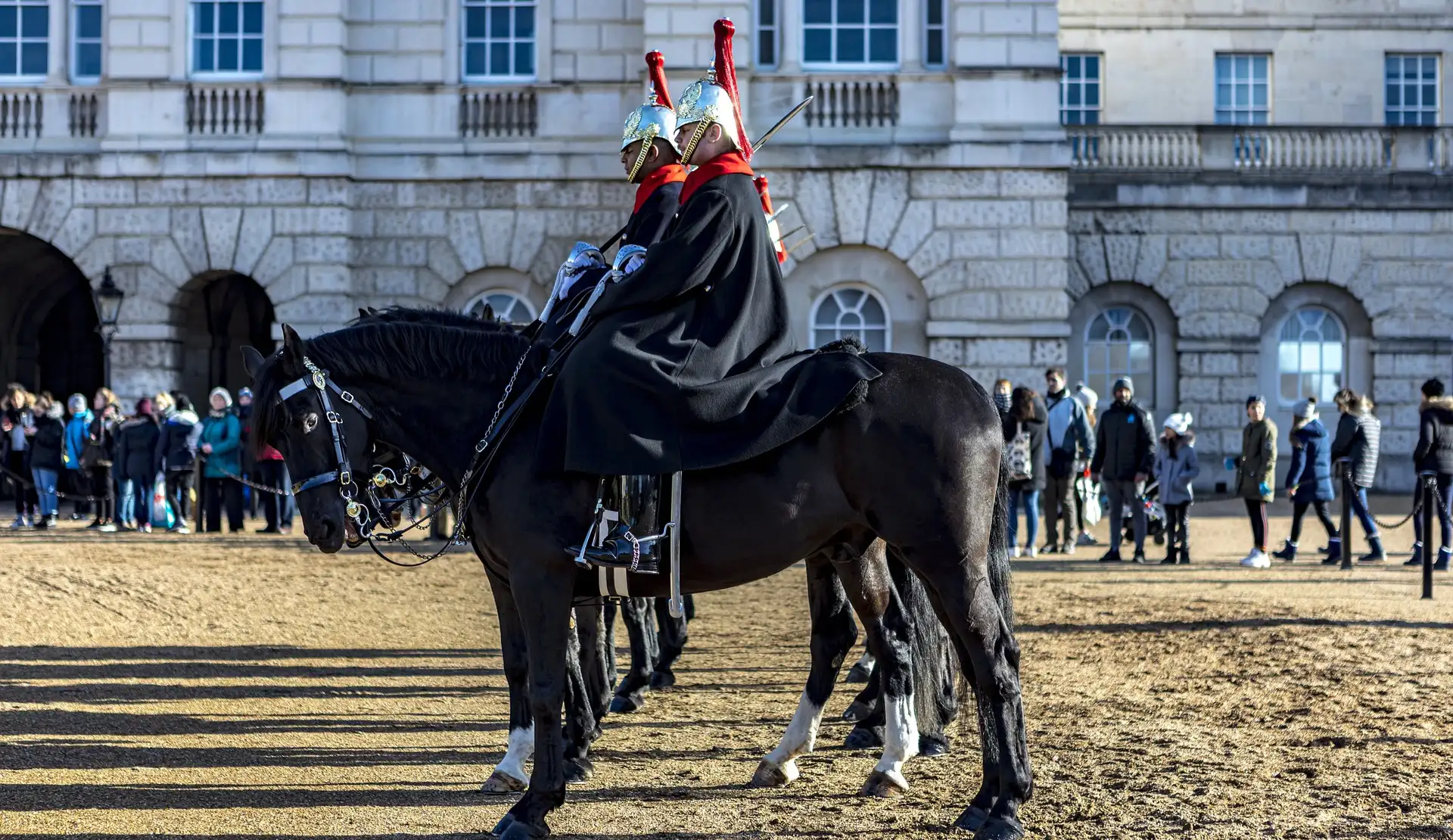 Zwei berittene Wachen mit glänzenden Helmen und schwarzen Umhängen auf schwarzen Pferden vor den Horse Guards in London, dahinter Besucher
