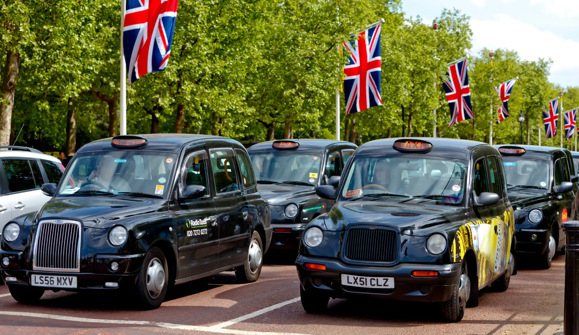 Mehrere schwarze Londoner Taxis stehen hintereinander auf einer von Bäumen gesäumten Straße, darüber hängen Union-Jack-Flaggen