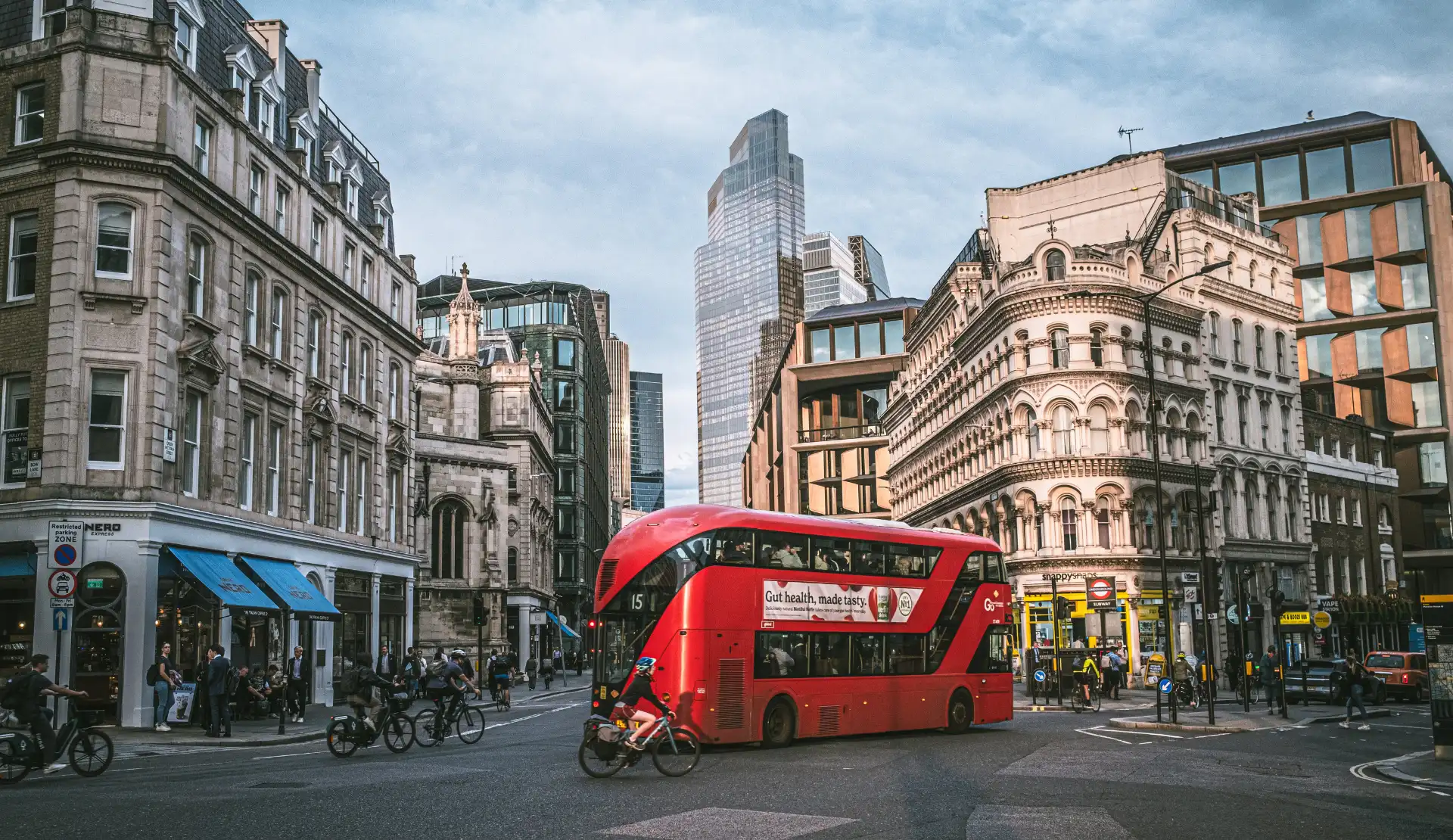 Roter Doppeldeckerbus fährt durch eine Straße in London, links und rechts historische Gebäude, im Hintergrund ein Hochhaus