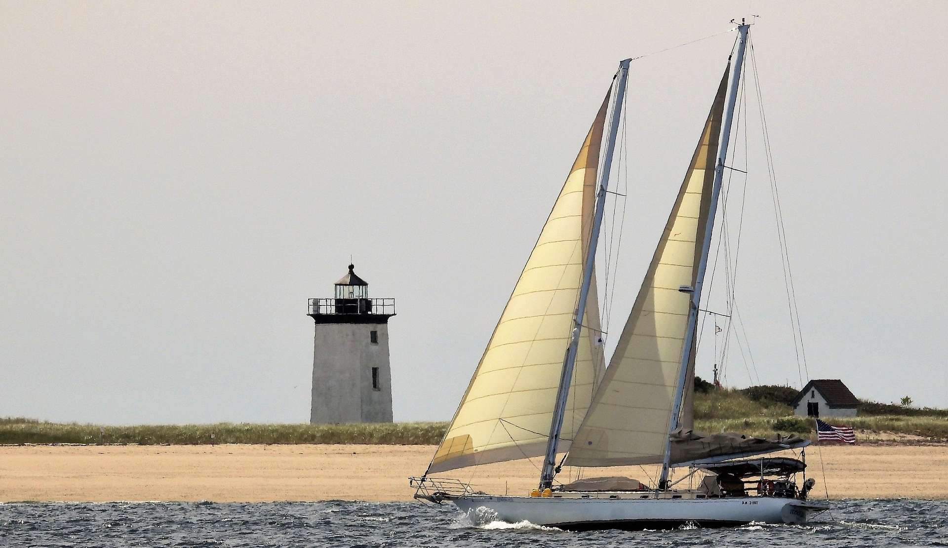 Segelboot mit zwei Segeln fährt nahe der Küste an einem Leuchtturm auf einer sandigen Landzunge vorbei.