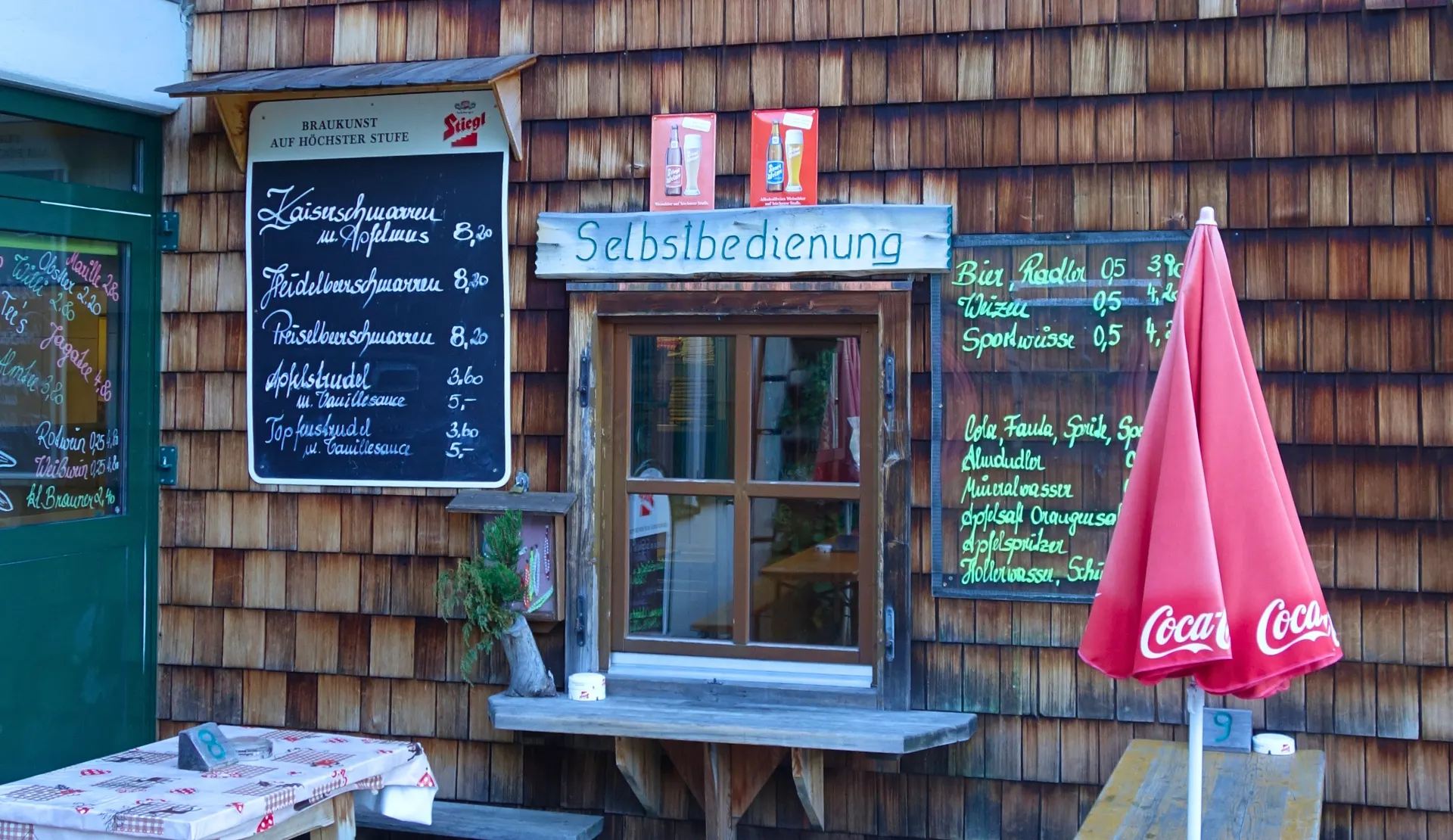 Selbstbedienung an einer Holzschindelwand mit Tafeln für Speisen und Getränke, daneben ein roter Sonnenschirm und ein Tisch auf der Terrasse der Tappenkarseehütte