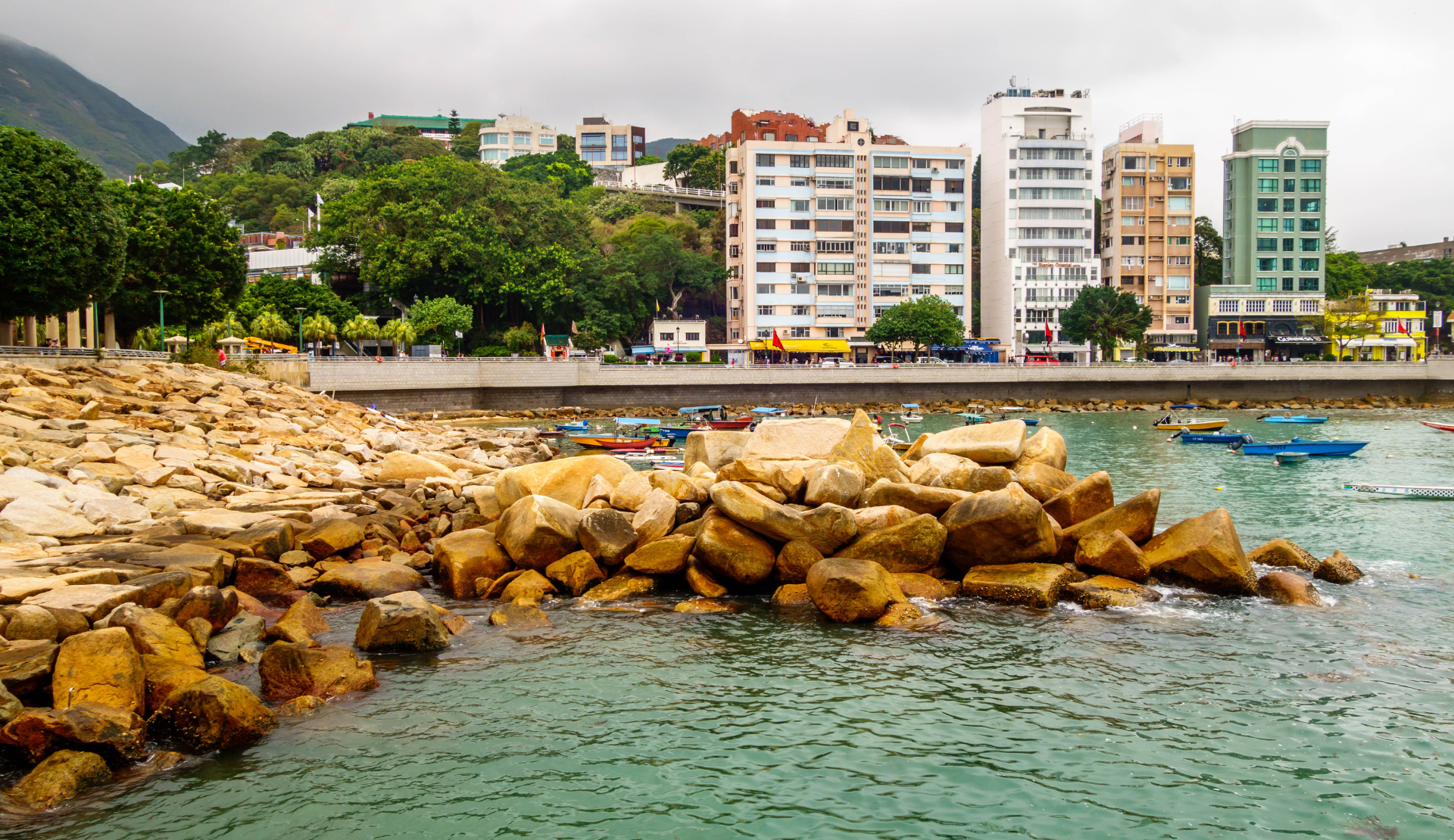 Stanley Waterfront in Hongkong mit goldenen Felsen im türkisfarbenen Wasser, bunten Wohnhäusern und grünen Bergen im Hintergrund