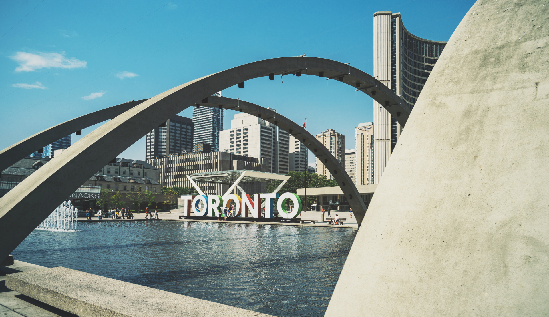 Blick auf das Toronto-Schild am Nathan Phillips Square mit Springbrunnen und moderner Skyline im Hintergrund.